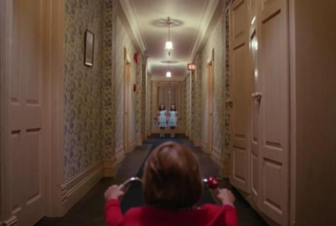 Child riding a bike in a hallway towards two girls in blue dresses.