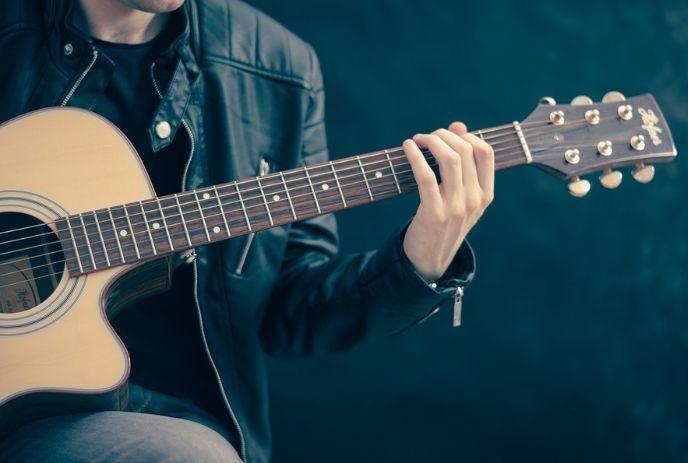 A musician in a black leather jacket playing a guitar in front of a dark background.