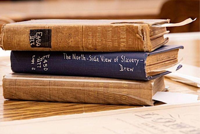 Three old books stacked on a wooden table. Titles visible: