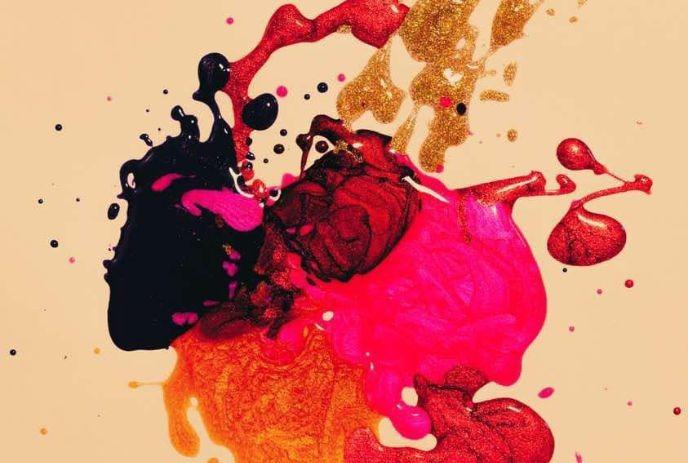 An abstract composition with splatters of black, red, pink, orange, and gold paint mixed on a light beige background.