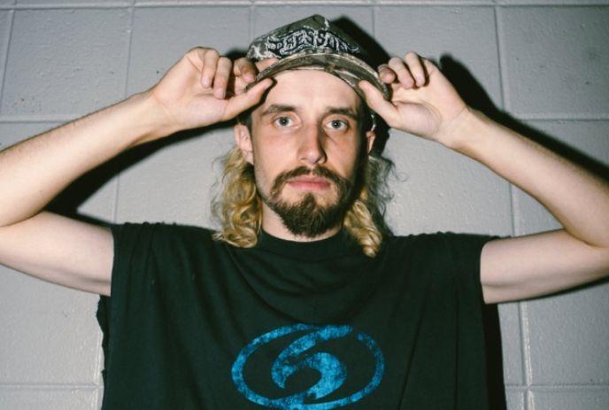 A person with long hair adjusting a patterned cap while wearing a black t-shirt with a blue circular logo.