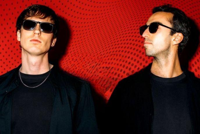 Two individuals stand in front of a red background with black dotted background. Wearing black clothing with black glasses.