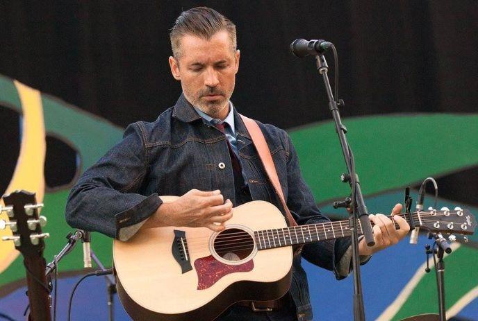 A person in a denim jacket plays an acoustic guitar on stage, with a microphone stand, colorful backdrop, and instruments.