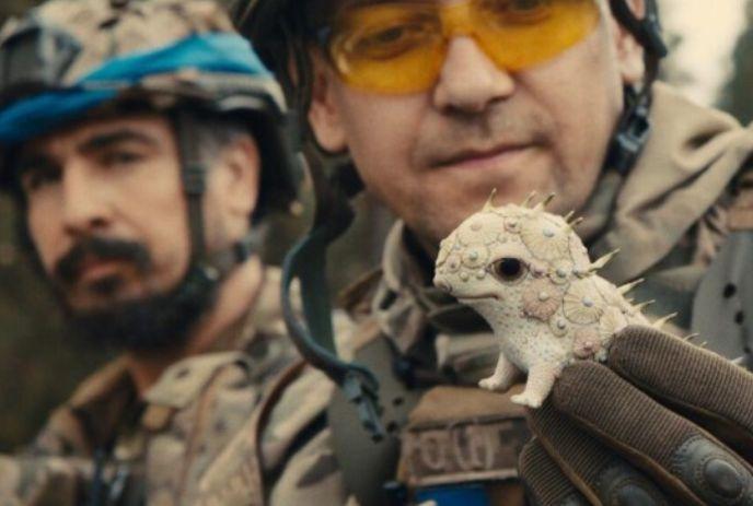 Two individuals in military uniforms hold a small, spiky lizard-like creature with pale, textured skin and small horns.