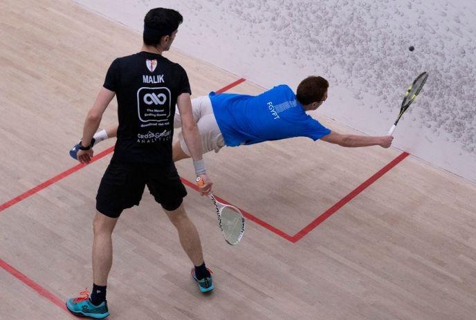 Two squash players in action, one lunging to hit the ball while the other watches.