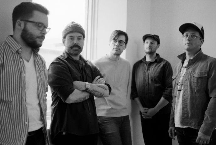 A black-and-white image of five men standing indoors, each wearing casual outfits posing.