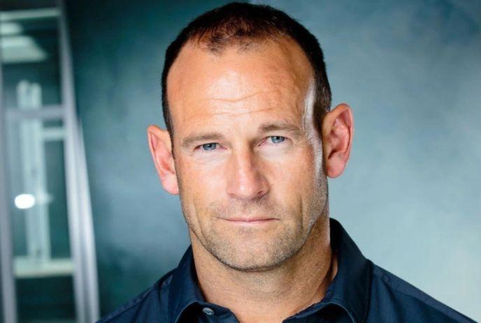 A headshot of David Nihill wearing a black shirt looking towards the camera under a dark theme background.