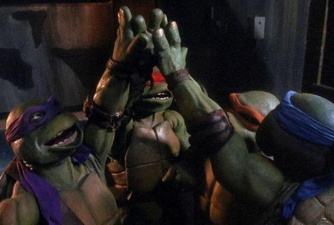 Three Teenage Mutant Ninja Turtles, each wearing colored masks (purple, red, blue), high fiving in a dimly lit setting.