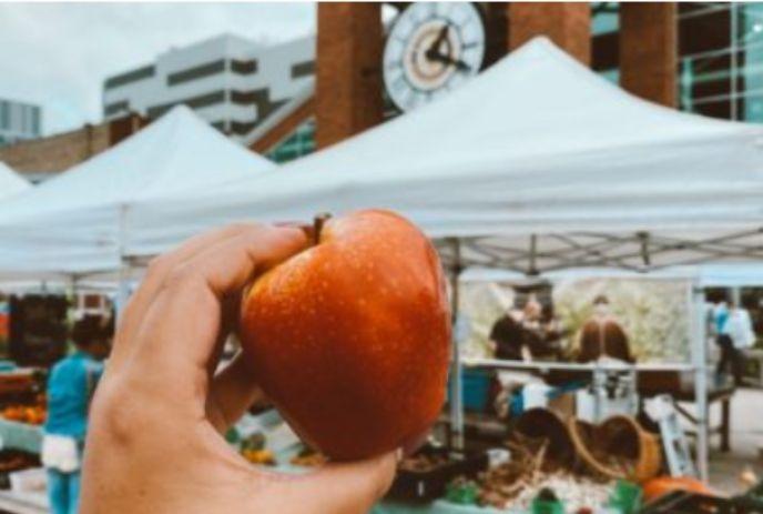 A hand holding a red apple in the foreground, with a market scene in the background featuring white tents and a clock tower.