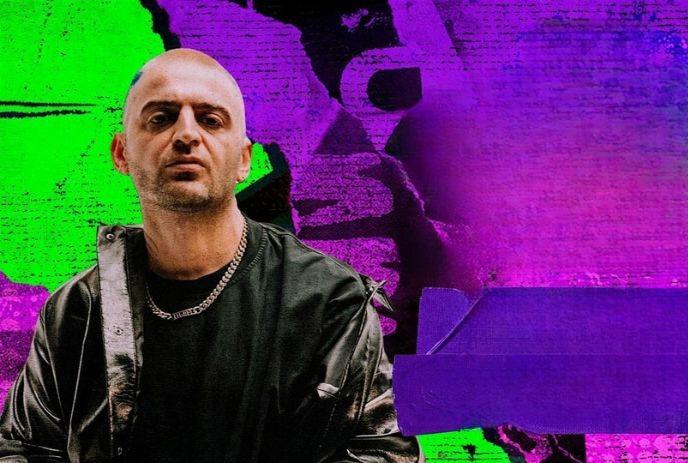 A man in a black jacket stands in front of a colorful, abstract background with green, purple, and pink hues.