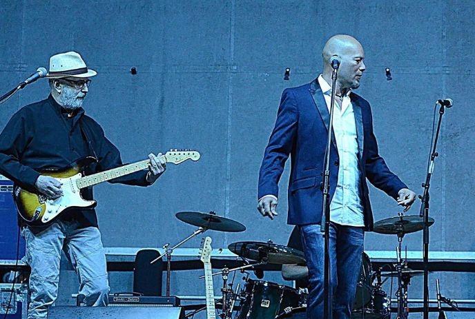 A guitarist on stage with blue lighting and a drummer in the background, creating an energetic concert atmosphere.