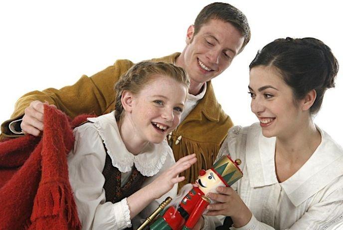 Three individuals, two of them hold a nutcracker doll while the person on the left wears a mustard-colored jacket