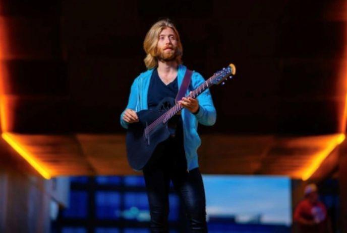 A person with a blurred face is playing a guitar in an urban setting with warm lighting and a blue sky in the background.