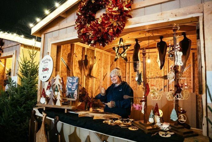 A wooden market stall adorned with a large red wreath showcases handcrafted items like ornaments, gnomes, and wooden figures