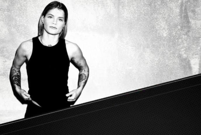 A woman with tattoos, in black tank top, posing for the picture with a black and white background setting.