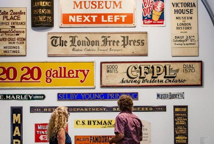Wall adorned with vintage signs, two individuals observing the collection from behind.