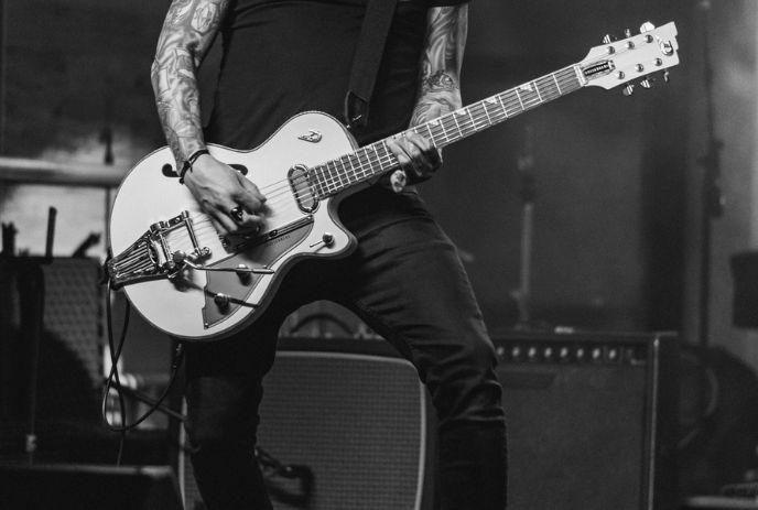 A black and white tattooed guitarist plays an electric guitar on stage and amplifier on the background