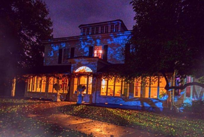 A two-story house at dusk with lights on, surrounded by trees, creating an eerie ambiance.