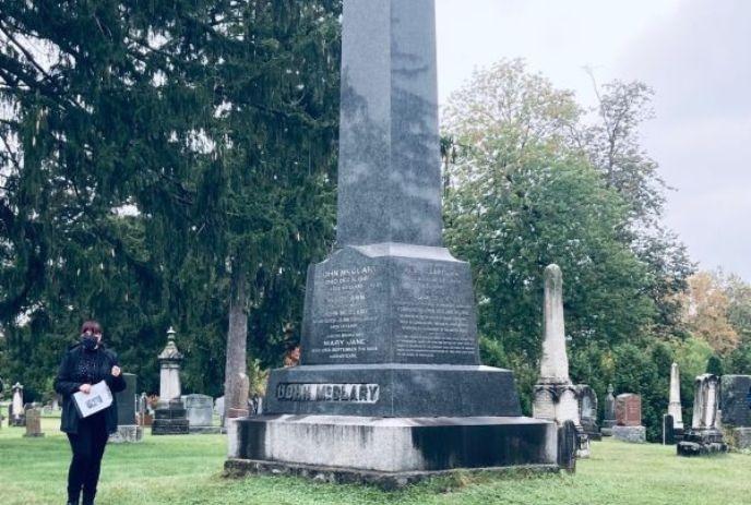 A woman holding papers, stands near a large monument in a cemetery with numerous headstones.