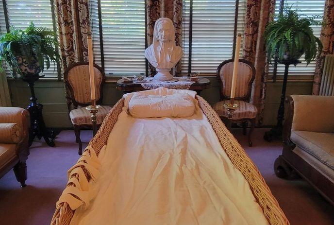 A wicker casket lies in an ornate room with elegant chairs, candles, and a bust statue. Ferns adorn the windowsill behind.