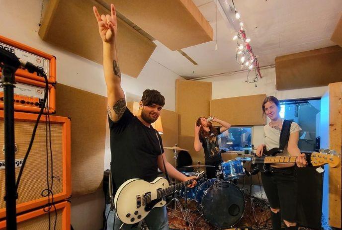 Three musicians in a soundproof room; one plays a white guitar; others play drums and bass guitar and posing for a picture.