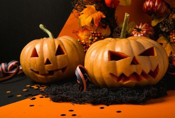 Two carved pumpkins with spooky faces on an orange backdrop, surrounded by autumn leaves and Halloween decorations.