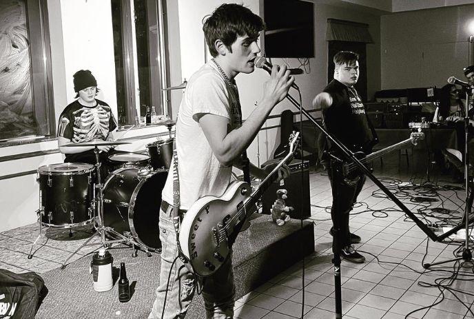 Black-and-white photo of a band on stage, guitarist singing, drummer and bassist playing in the background.