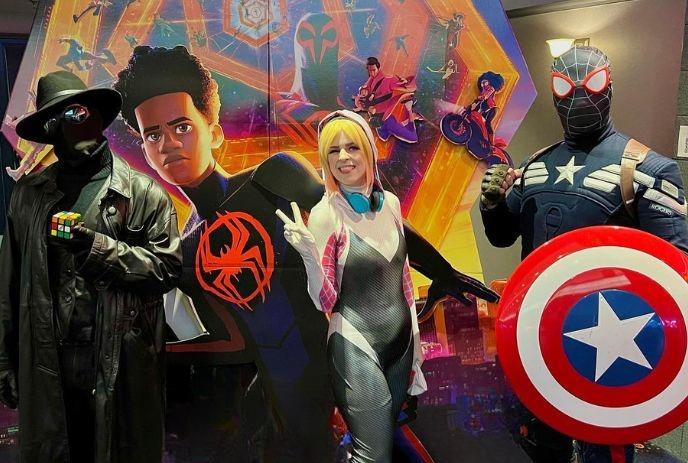 Three artists in superhero costumes pose in front of a colorful Spider-Man-themed backdrop.