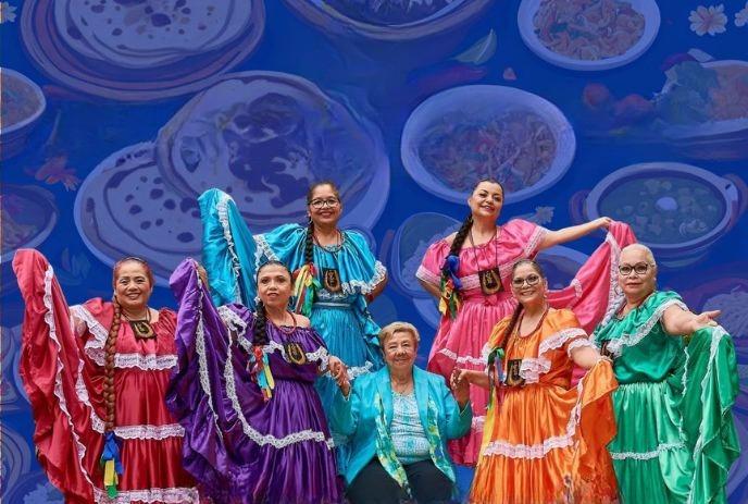 A group of performers in their cultural costumes posing with pupusas in the blue shady background.