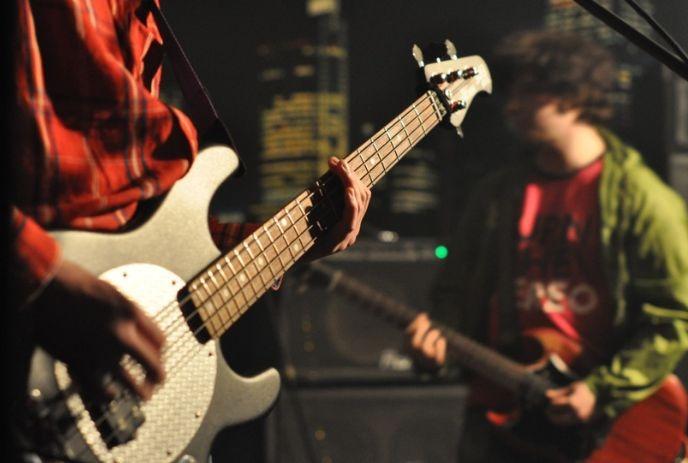 A person plays an electric guitar, while in the background, a second guitarist is blurred in motion,