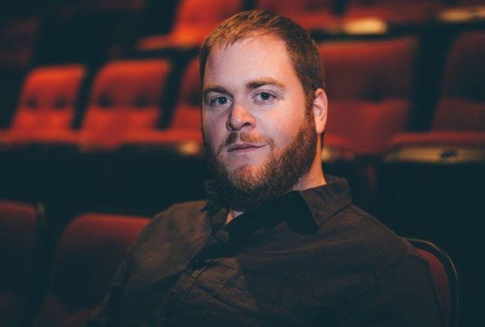 A man with a trimmed beard and short hair sits in an empty theater, wearing a dark shirt, looking calmly at the camera.