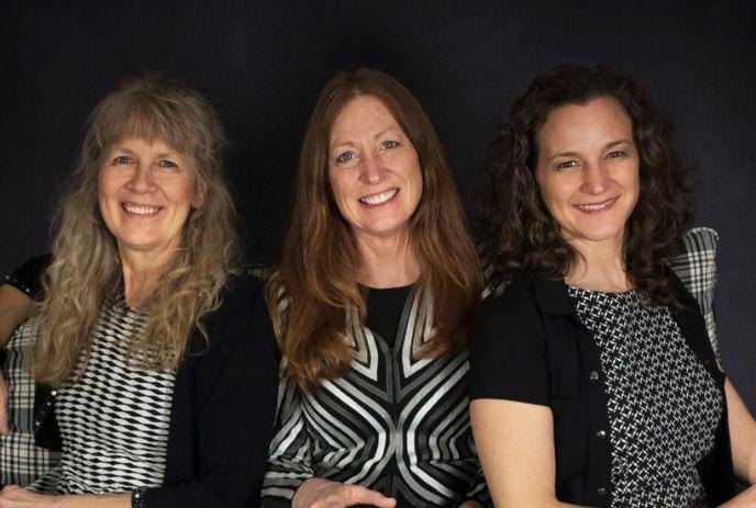 Three women artists sitting together and smiling, posing for a picture under a dark black background.