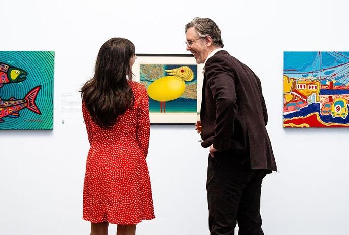 Two people viewing colorful paintings in an art gallery under a white background.