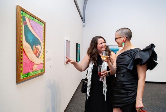 Two women in black outfits in a gallery observe a colorful painting under a white background.