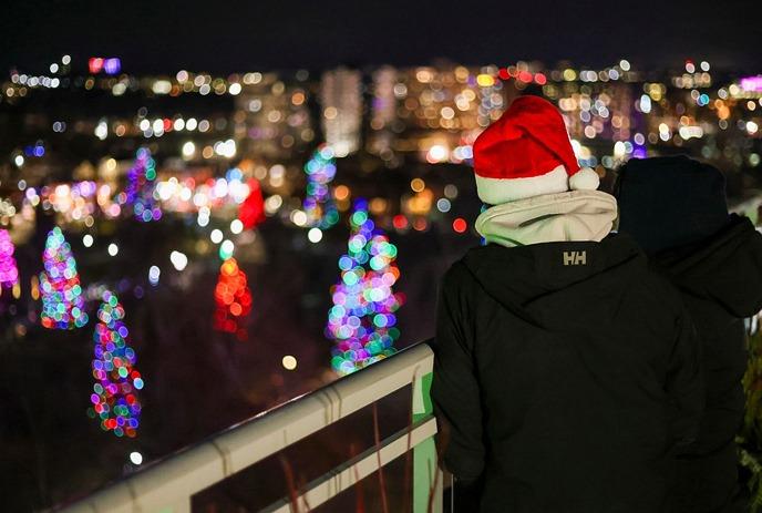 Two people stand on a balcony overlooking a cityscape at night, with streets and trees adorned with colorful holiday lights