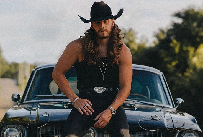 Warren Zeiders poses confidently on the hood of a classic black car against a backdrop of trees and power lines.