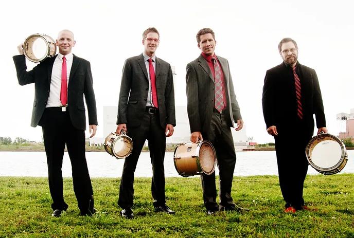 A group of five musicians playing various percussion instruments including congas, bongos, marimbas, and xylophones.