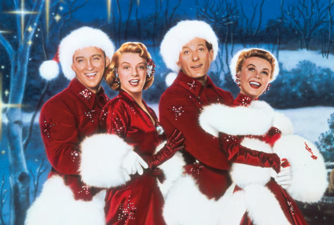 People in festive red and white Christmas attire stand together in a snowy nighttime scene with trees in the background.