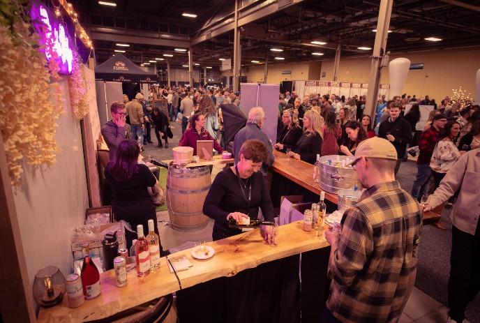 Attendees gathered at the London Wine & Food Show, enjoying wine and food from various booths and a wooden bar setup.