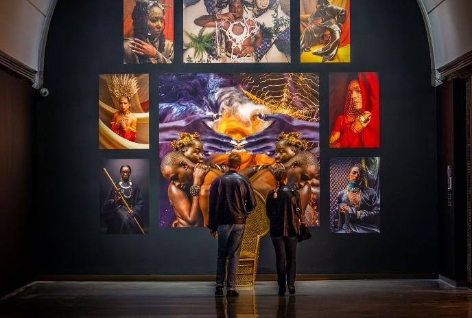 Two people stand in front of a vibrant wall of Afrofuturistic art, admiring colorful, imaginative portraits.