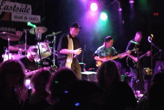 A group of five artists stands together in a dimly lit room. There's a red fire extinguisher on the wall behind them.