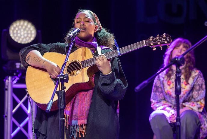 A female musician singing and playing an acoustic guitar live on stage with a backup singer in the background