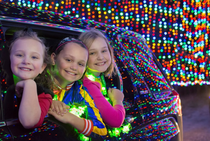 Three children with faces blurred, leaning out of a car window, surrounded by colorful holiday lights.