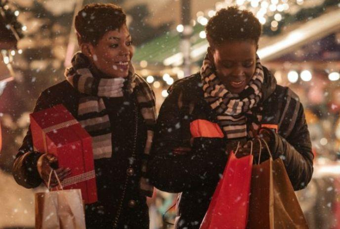 Two people wearing scarves and jackets are holding shopping bags and a gift box while walking in a festive.