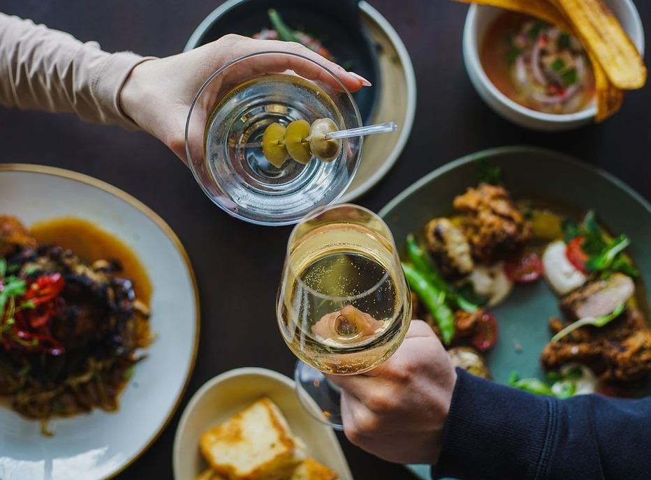 Two hands holding a martini and the other a glass of wine on opposite ends of a table with various plates of food