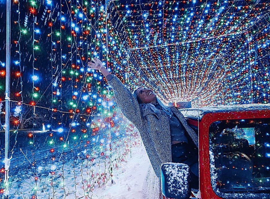 A female stretching out from the passenger side of a vehicle while its driving through a tunnel of coloured Christmas lights